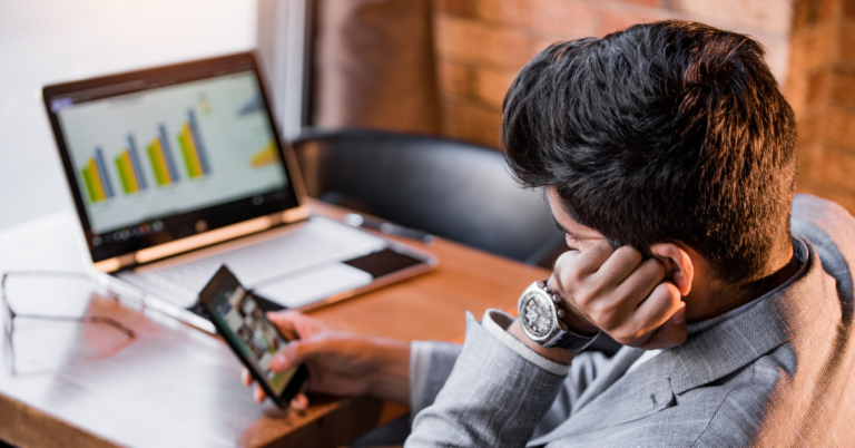 Um homem na frente do notebook mexendo no celular e deixando o trabalho de lado, usando a procrastinação